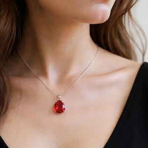 Puede incluir: Un collar de plata con un gran colgante de piedra roja en forma de lágrima. El colgante está engastado en un marco de plata y cuelga de una delicada cadena de plata. El collar se lleva sobre una blusa negra.