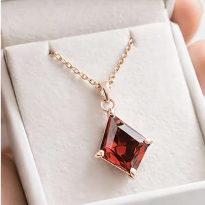 May include: A rose gold necklace featuring a large, square-cut, deep red gemstone pendant. The pendant is set in a four-prong setting and hangs from a delicate chain. The necklace is displayed in a white jewellery box.