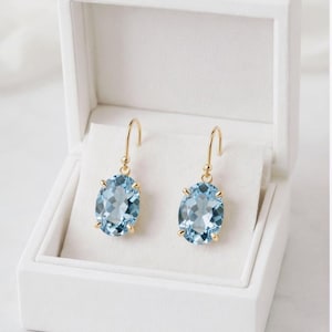 Puede incluir: Un par de pendientes colgantes dorados con gemas ovaladas de color azul claro. Los pendientes se exhiben en una caja de joyería blanca abierta con un interior suave y blanco. Las gemas están facetadas.