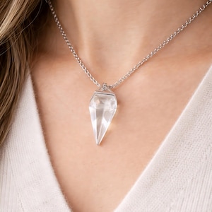 Peut inclure: Un collier avec un pendentif en cristal clair et une chaîne argentée. Le cristal a une forme géométrique pointue. Le collier est porté sur un pull blanc.