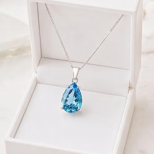 Può includere: Una collana d'argento con un grande pendente a forma di pera, in pietra preziosa blu chiaro. Il pendente è incastonato in una lunetta d'argento e pende da una delicata catena d'argento. La collana è esposta in un portagioie bianco aperto.