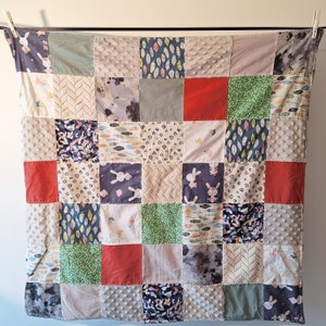 Peut inclure: Une courtepointe patchwork avec une variété de tissus dans un motif carré. La courtepointe présente des carrés de tissus rouges, verts, bleus et gris, certains avec des motifs comme des lapins, des plumes et des points texturés. La courtepointe est suspendue à une corde à linge.