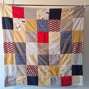 Peut inclure: Une courtepointe patchwork avec une variété de carrés de tissu rouges, marine, jaunes, gris et blancs. Certains carrés présentent des motifs tels que des fusées, des pois et des chevrons. La courtepointe est suspendue à une corde à linge.