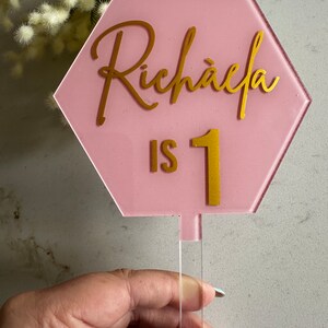 以下が含まれることがあります： ピンクの六角形のケーキトッパーに、金色の文字で「Richaela is 1」と書いてあります。