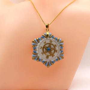Peut inclure: Un collier à chaîne dorée avec un pendentif hexagonal au centre blanc, un symbole pentagramme doré et de petites phases lunaires. Le bord du pendentif est orné de formes en pétale bleu clair et dorées alternées et d'une petite perle bleue.