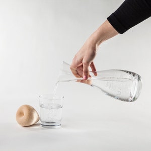 Könnte beinhalten: Eine klare Glaskaraffe gießt Wasser in ein klares Glas. Ein holzfarbenes, donut-förmiges Objekt liegt links vom Glas. Die Karaffe hat ein geschwungenes Design und wird von einer Person mit einem schwarzen Langarmshirt gehalten.