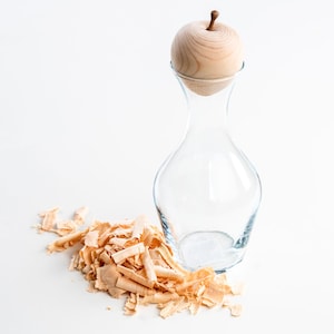 Könnte beinhalten: Eine klare Glasflasche mit einem Stopfen in Form eines Holzapfels. Die Flasche steht auf einer weißen Oberfläche mit Holzspänen, die um sie herum verstreut sind.