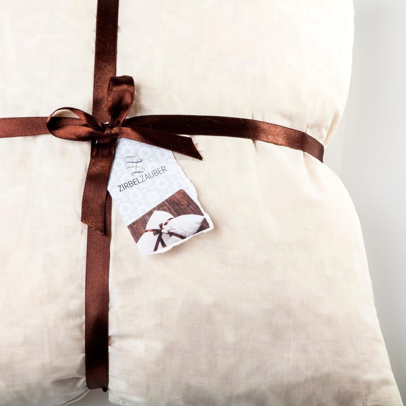 May include: A white pillow with a brown ribbon tied around it. The ribbon has a tag attached that says "Zirbelzauber".