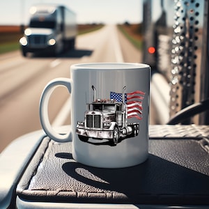 May include: White ceramic mug with a graphic of a semi-truck with an American flag waving behind it. The truck is gray and white with a black grill. The flag is red, white, and blue.