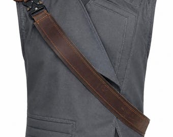 May include: A charcoal gray sleeveless vest featuring a high collar, a diagonal front overlap, and a brown leather-like shoulder strap secured with a black metal buckle. The garment includes stitched panel detailing and a side buckle adjustment strap.