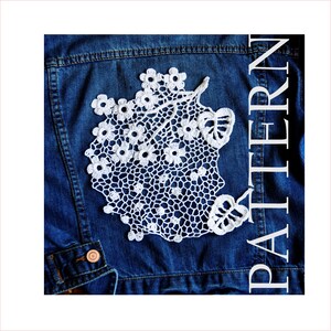 May include: A white, crocheted floral applique is displayed on a blue denim jacket. The applique features a delicate openwork design with small flower and leaf details. The word "PATTERN" is printed vertically on the right side of the image.