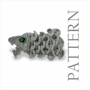May include: A crocheted gray dragon with a green eye. The dragon has a detailed scale pattern and an open mouth. The word "PATTERN" is printed vertically on the right side of the image.