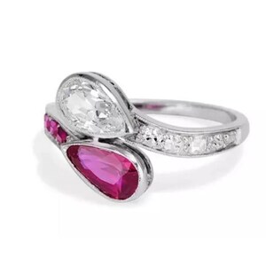 Puede incluir: Un anillo de plata con una piedra preciosa blanca en forma de pera y una piedra preciosa de rubí en forma de pera. El anillo también tiene pequeñas piedras preciosas redondas y transparentes a lo largo de la banda. Sobre fondo blanco.