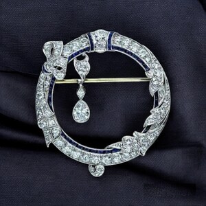 May include: A circular brooch featuring a silver-toned frame encrusted with numerous clear white diamonds and accented by thin strips of deep blue sapphires. A single pear-shaped diamond pendant hangs from the top inner edge of the circle.