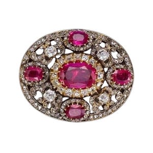 Könnte beinhalten: Ovale Brosche mit einem zentralen Rubin, umgeben von kleineren Rubinen und Diamanten. Die Brosche hat eine Gold- und Silberfassung mit aufwendigen Filigranarbeiten. Die Edelsteine sind in einem symmetrischen Muster angeordnet.