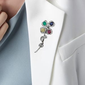 May include: A floral brooch with four gemstone petals in green, blue, yellow, and red, each surrounded by small clear stones. The brooch is attached to a white blazer, with a light blue shirt visible underneath.