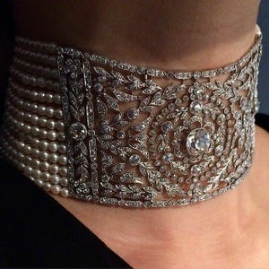 May include: An ornate choker necklace featuring multiple strands of pearls and a wide, detailed silver band. The band is embellished with intricate floral and leaf patterns, accented with sparkling gemstones, and a large central diamond.