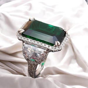 May include: A silver ring featuring a large emerald green gemstone encircled by smaller clear gemstones. The ring has a smaller clear gemstone on each side of the central stone.
