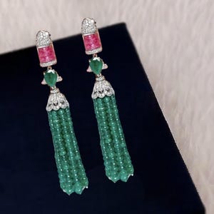Puede incluir: Un par de pendientes de borla de piedra preciosa verde con acentos rosa y blanco. Los pendientes presentan una gota de piedra preciosa verde, una parte superior de piedra preciosa rosa y blanca y una larga borla verde.