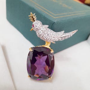 Könnte beinhalten: Ein Vogelbrosche mit goldener Krone und Füßen, die auf einem großen, kissenförmigen, lila Edelstein sitzt. Der Körper des Vogels ist mit funkelnden weißen Steinen besetzt und hat ein kleines rotes Auge.
