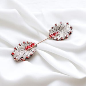 Puede incluir: Un broche de plata con dos piezas en forma de abanico, cada una adornada con piedras preciosas rojas y un diseño de filigrana de plata.