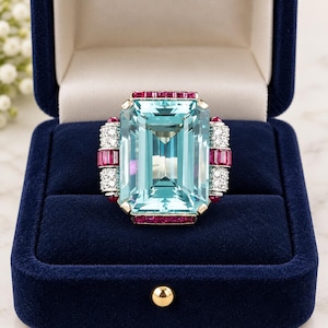 Könnte beinhalten: Ein Ring mit einem großen, smaragdgeschliffenen hellblauen Edelstein als Mittelstück, flankiert von vertikalen Reihen kleiner weißer Diamanten und rechteckiger roter Rubine. Die Fassung ist metallisch und der Ring liegt in einer blauen Samtschatulle.