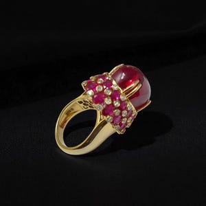 May include: A luxurious gold ring featuring a large, round, red gemstone as the centerpiece. The ring is adorned with smaller red and clear gemstones, creating a striking contrast against the gold band. The ring is set against a black background.