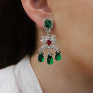 Op de afbeelding: Een elegante oorbel met een grote ovale groene edelsteen aan de bovenkant, een bloemmotief met een centrale rode edelsteen en drie druppelvormige groene edelstenen. De oorbel is versierd met kleine, sprankelende, heldere edelstenen.