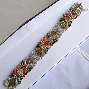 Könnte beinhalten: Ein Gliederarmband mit Blumenmotiven, besetzt mit gelben, roten und violetten Edelsteinen, akzentuiert durch grüne Emaille-Blätter und klare Pavé-Steine. Das Design umfasst silberfarbene Glieder und einen sicheren Verschlussmechanismus am Ende.