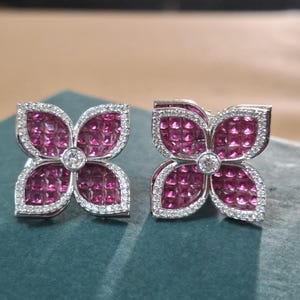 Könnte beinhalten: Ein Paar blumenförmige Ohrringe aus silberfarbenem Metall mit vier Blütenblättern, die mit quadratisch geschliffenen magentafarbenen Edelsteinen besetzt sind. Jedes Blütenblatt ist von kleinen, klaren Pavé-Steinen umrandet, mit einem runden Stein in der Mitte.