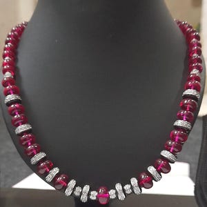 Pode incluir: Um colar de contas vermelho rubi com detalhes prateados e pedras transparentes. O colar &eacute; exibido em um manequim preto. As contas s&atilde;o de cor vermelha profunda, e os detalhes prateados s&atilde;o intercalados ao longo do colar.