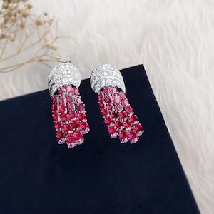 May include: A pair of silver-toned earrings with a curved top set with small, clear stones. Below, strands of bright red gemstones hang in a tassel design. The earrings are presented on a black surface, set against a fluffy white background.