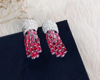 Puede incluir: Un par de pendientes elegantes con una parte superior plateada curvada incrustada con pequeñas piedras transparentes. Debajo, hileras de gemas rojas vibrantes crean un efecto de borla. Los pendientes se exhiben sobre una superficie negra con un fondo blanco y esponjoso.