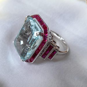 May include: A silver ring featuring a large, rectangular, light blue faceted gemstone as the centerpiece. The stone is surrounded by a border of small, square-cut, deep red rubies. The metal band has intricate filigree detailing on the sides.