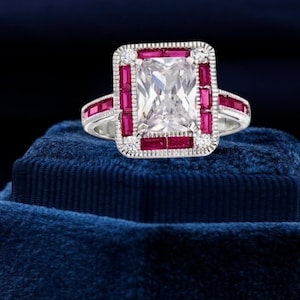 Puede incluir: Un anillo de plata con una gran piedra preciosa rectangular transparente rodeada por un borde de piedras rojas más pequeñas. El anillo está sobre una caja de terciopelo azul.