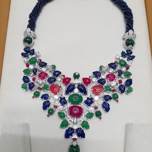 Może przedstawiać: Ozdobny naszyjnik z ciemnoniebieskim kołnierzem z koralików i wisiorkiem o kwiatowym wzorze. Wisiorek zdobiony jest czerwonymi, zielonymi, niebieskimi i białymi kamieniami szlachetnymi. Naszyjnik ma centralną zieloną kroplę z kamienia szlachetnego.