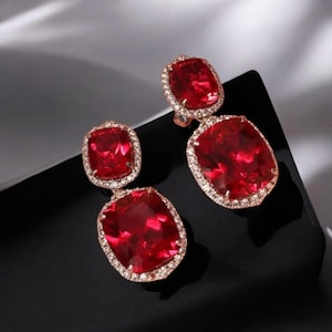 May include: Rose gold earrings with striking red gemstones. Each earring has a rectangular top stone and a larger, oval-shaped bottom stone, both encircled by a halo of small, clear stones. The earrings are presented on a black surface, creating a contrast.