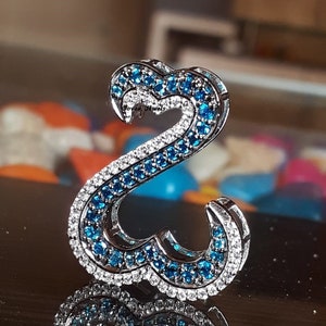 May include: A silver-toned pendant in the shape of a stylized letter "S". The pendant is encrusted with small, round, blue and white gemstones. The gemstones are arranged in a pattern that highlights the curves of the letter. The pendant is highly reflective.