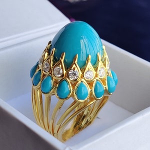Puede incluir: Un anillo de oro con una gran piedra turquesa en cabujón rodeada de un halo de piedras turquesas más pequeñas y diamantes.