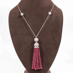 Puede incluir: Un collar de plata con un colgante de borla. El collar presenta tres acentos redondos y brillantes y una larga borla hecha de pequeñas cuentas rojas. El colgante tiene una parte superior decorativa y ornamentada. El collar se exhibe en un busto de joyería marrón.