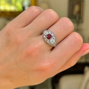 Puede incluir: Anillo de estilo vintage con un rubí central ovalado y dos piedras preciosas redondas y claras. El anillo está engastado en un metal plateado con intrincados detalles alrededor de las piedras.