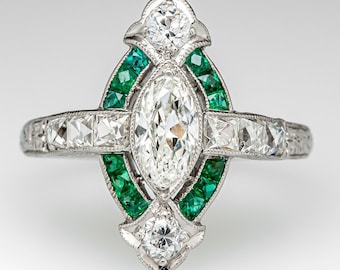 May include: A vintage engagement ring with a marquise-shaped diamond surrounded by a halo of emerald green stones. The ring is set in platinum and features a delicate filigree design.