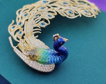 Peut inclure: Une broche en or en forme de paon avec émail bleu et vert et strass transparents. Les plumes de la queue du paon sont déployées derrière lui.