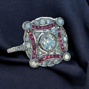 May include: A silver ring with a large round diamond surrounded by smaller diamonds and red rubies. The ring has a square shape and is set in a geometric design.