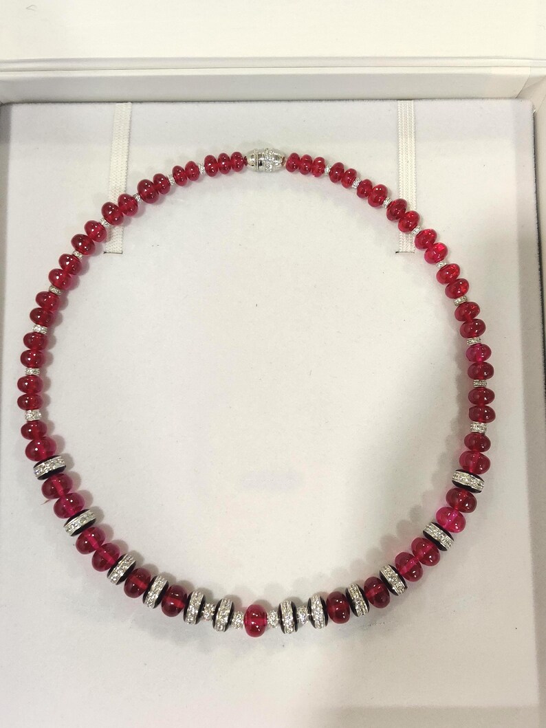 Pode incluir: Um colar de contas vermelhas com detalhes em prata e diamantes. O colar &eacute; composto por contas vermelhas polidas, com se&ccedil;&otilde;es de espa&ccedil;adores prateados cravejados com diamantes. O fecho &eacute; prateado e tem um design de diamante.