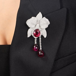 Könnte beinhalten: Eine Brosche in Form einer weißen Diamant-Orchidee mit einem großen roten Edelstein in der Mitte und zwei kleineren roten Edelsteintropfen, die unten hängen.