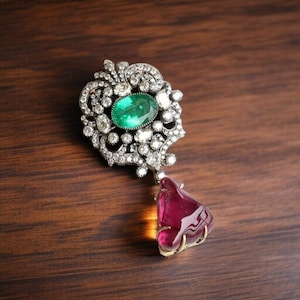May include: An elaborate pendant with an oval emerald centre, encircled by small diamonds. A teardrop-shaped, deep red gemstone hangs below. The pendant has a silver-coloured metal setting, displayed on a dark wood surface.