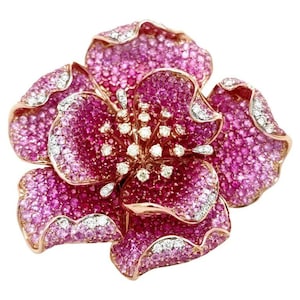 Op de afbeelding: Een bloemvormige broche van roségoud met gelaagde bloemblaadjes die dicht bezet zijn met levendige roze saffieren. De randen van de bloemblaadjes zijn geaccentueerd met kleine, heldere witte diamanten en het midden toont een cluster van ronde witte diamanten.
