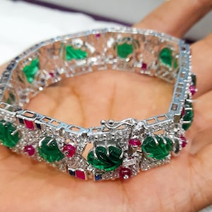 May include: A silver bracelet with emerald green leaf-shaped stones, small red and blue gemstones, and clear crystals. The bracelet is set with a clasp and is displayed on a hand.