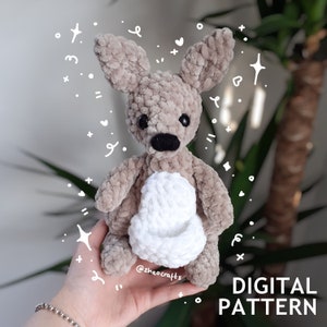 May include: A crocheted kangaroo plush toy with a white pouch. The toy is made with a fuzzy yarn and has black eyes and a black nose. The toy is being held in a hand with a silver bracelet.